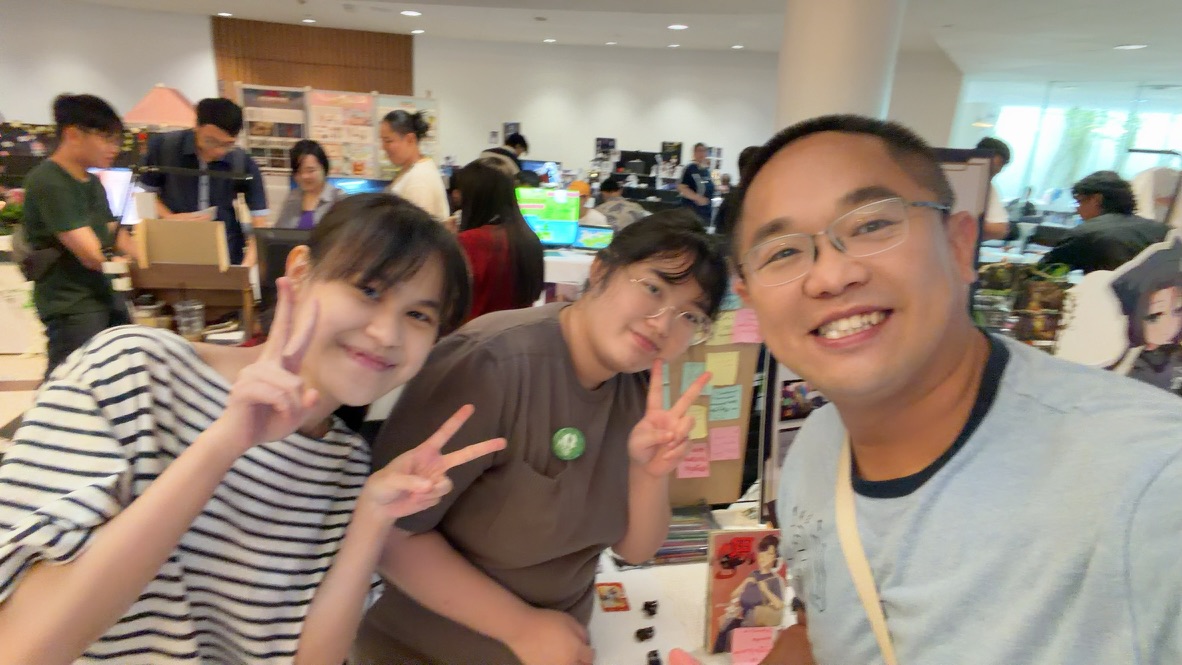 Terry posing with two smiling artists among busy illustration booths in Bangkok