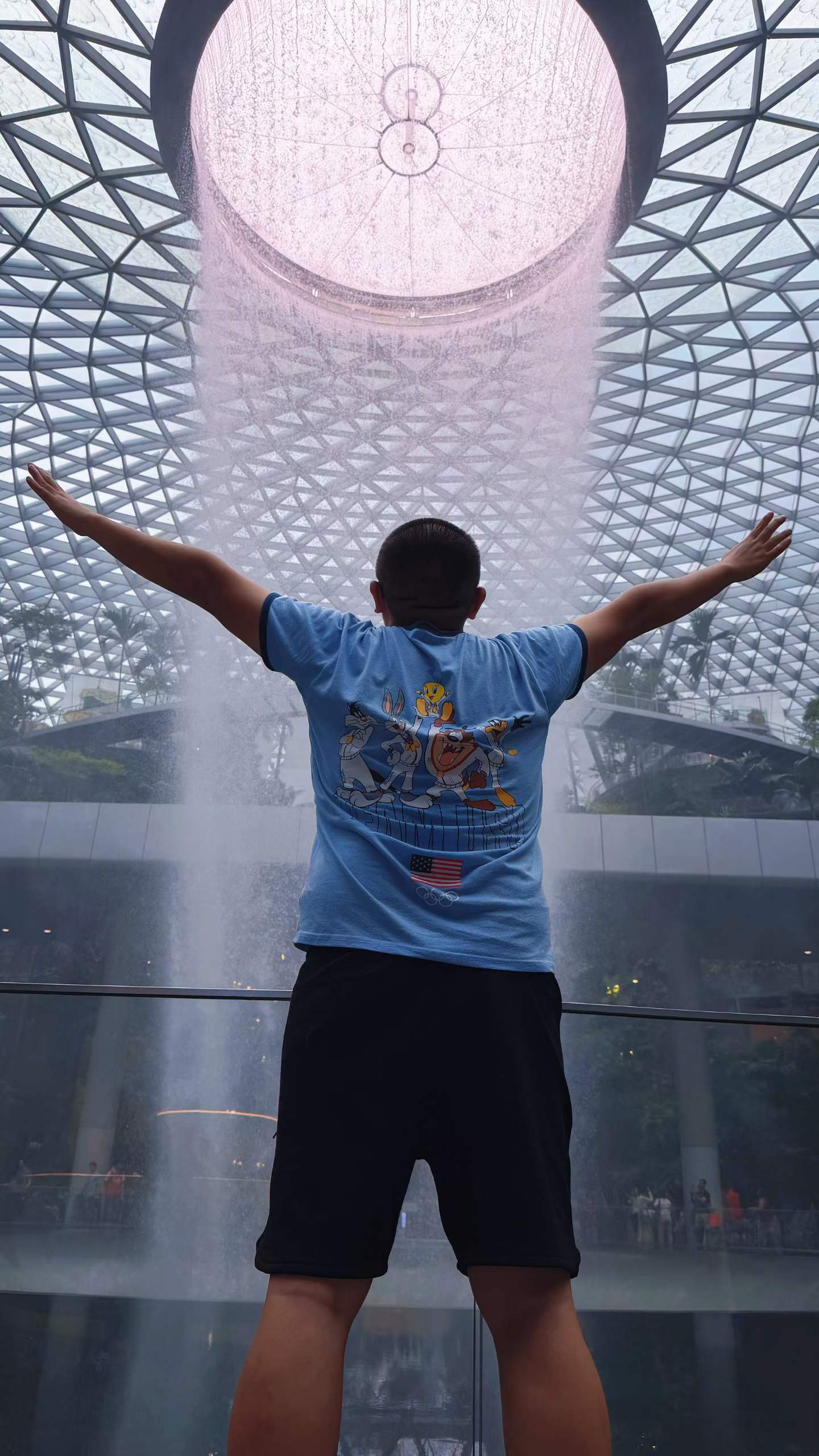 Terry standing with arms outstretched in front of the Rain Vortex waterfall at Jewel Changi Airport
